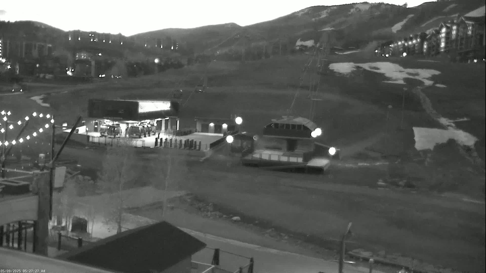 A nighttime, grayscale image shows a ski lift and buildings near a mountain slope with patches of snow under a dark sky.