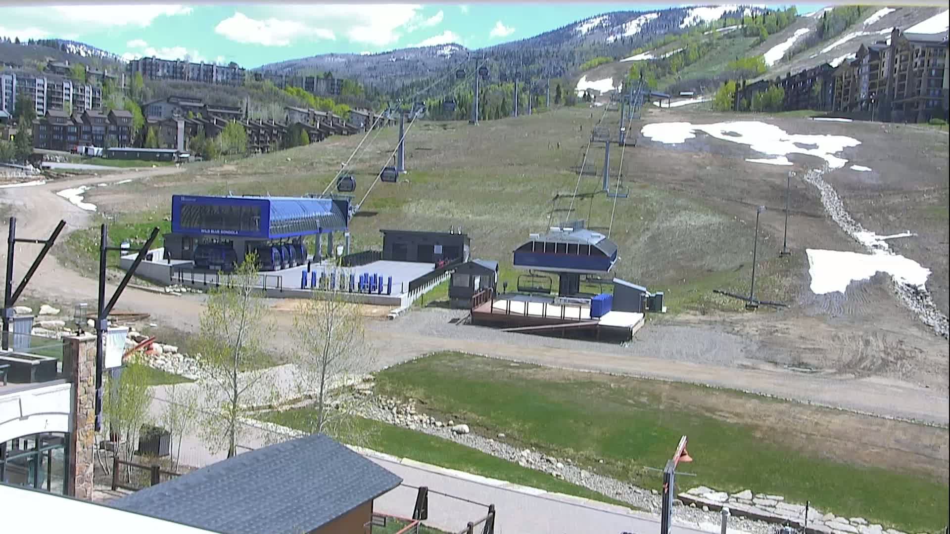 A mostly sunny day reveals a ski resort base area with two chairlifts, some buildings, and patches of remaining snow on the mountainside.