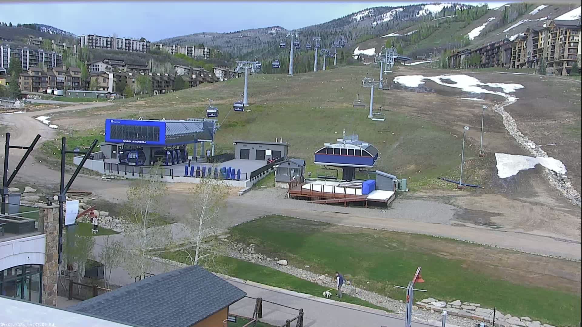 A mostly barren ski slope with several chairlifts and a gondola station, surrounded by buildings and some patchy snow, under a partly cloudy sky.