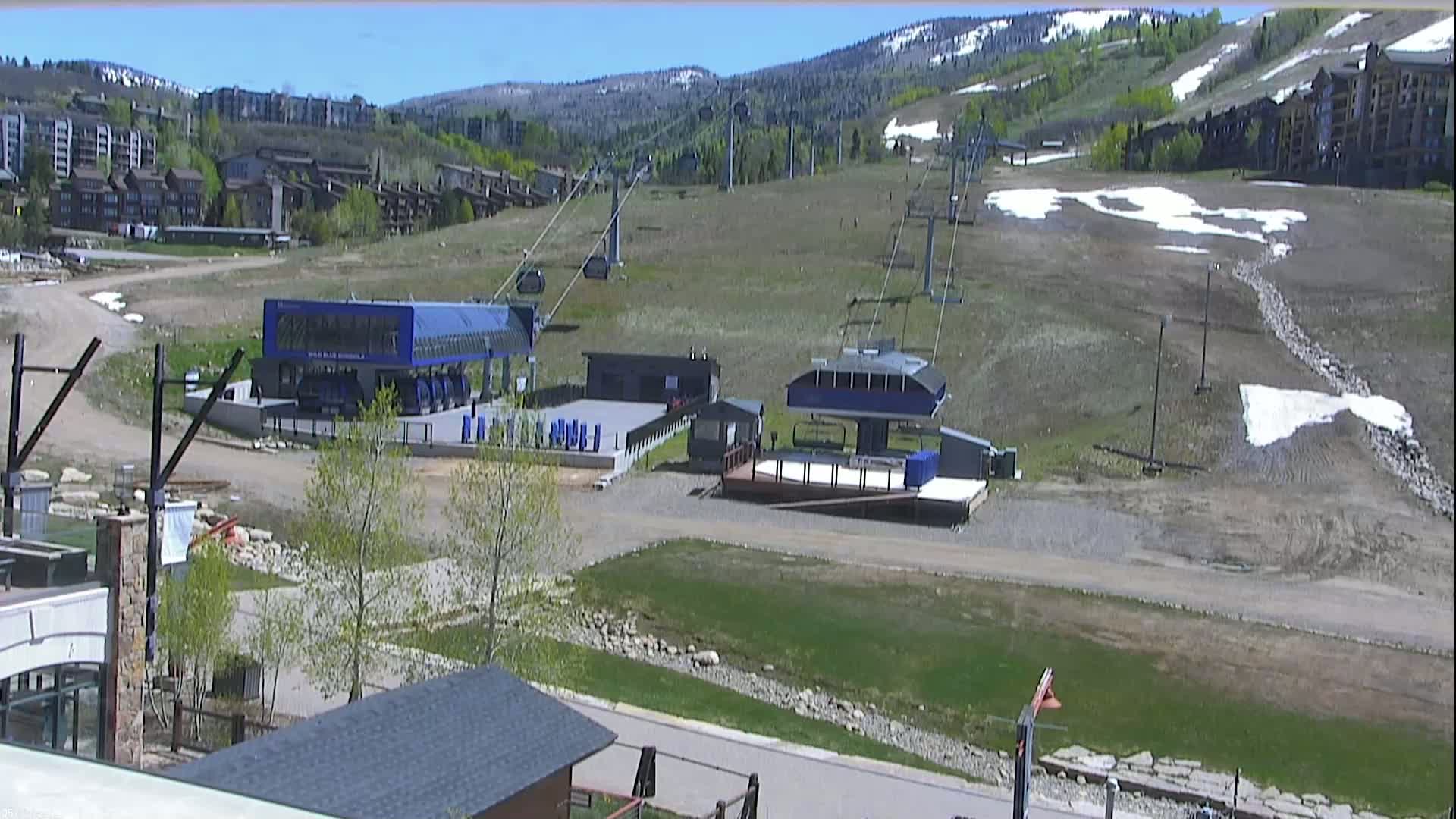 A mostly sunny day shows a ski resort with two chairlifts and their base buildings, surrounded by brown grassy hills with patches of snow, and buildings in the background.