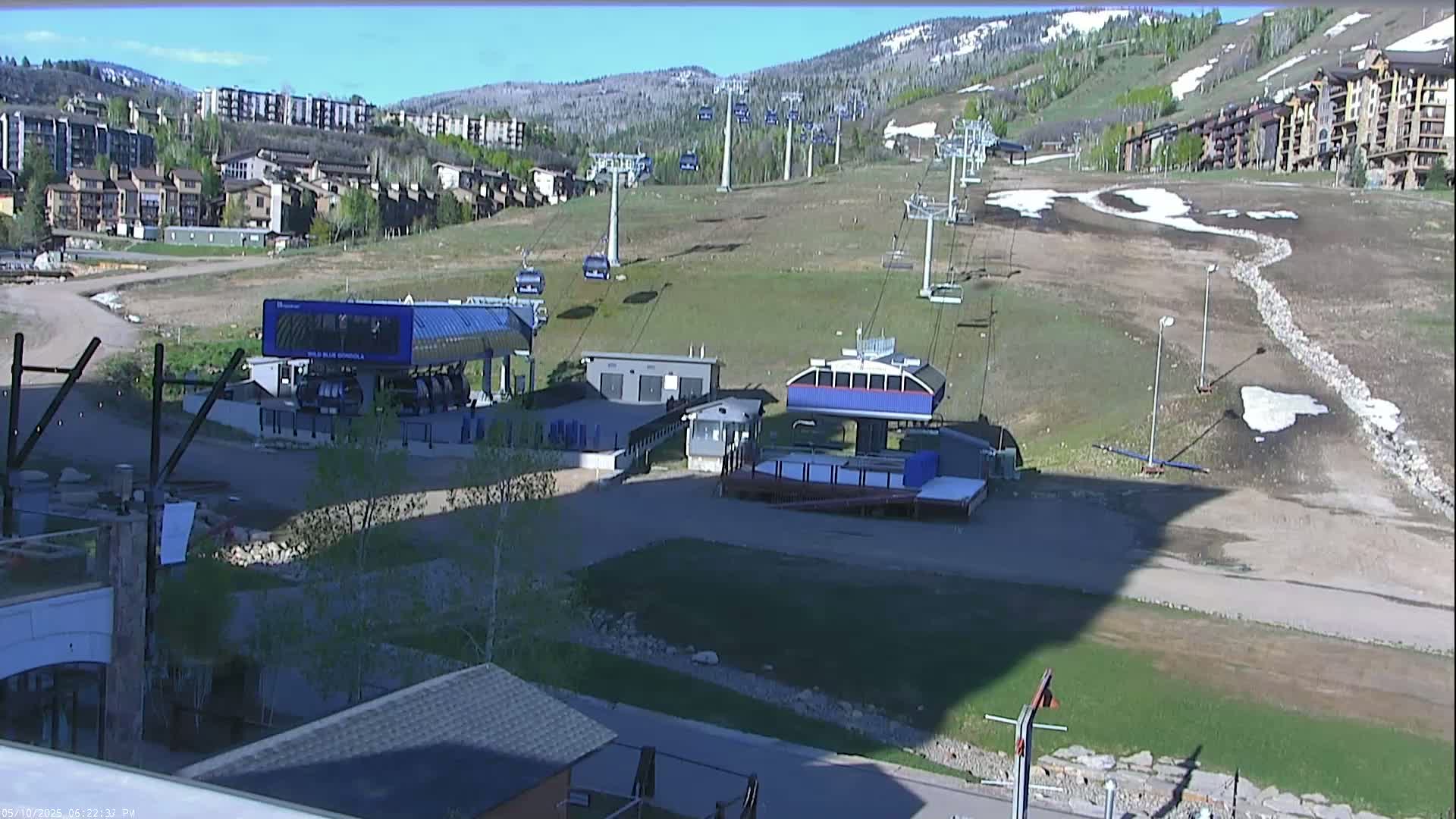 A mostly sunny view of a ski resort in spring, showing chairlifts, buildings, and patches of melting snow on a grassy hill.