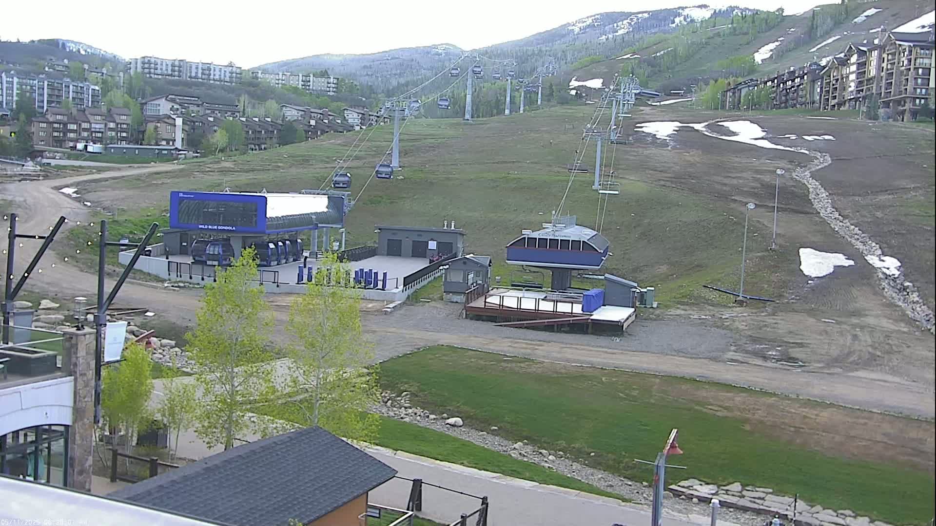 A ski resort base area with chairlifts and gondolas, showing patchy snow and early spring vegetation on the mountainside under an overcast sky.