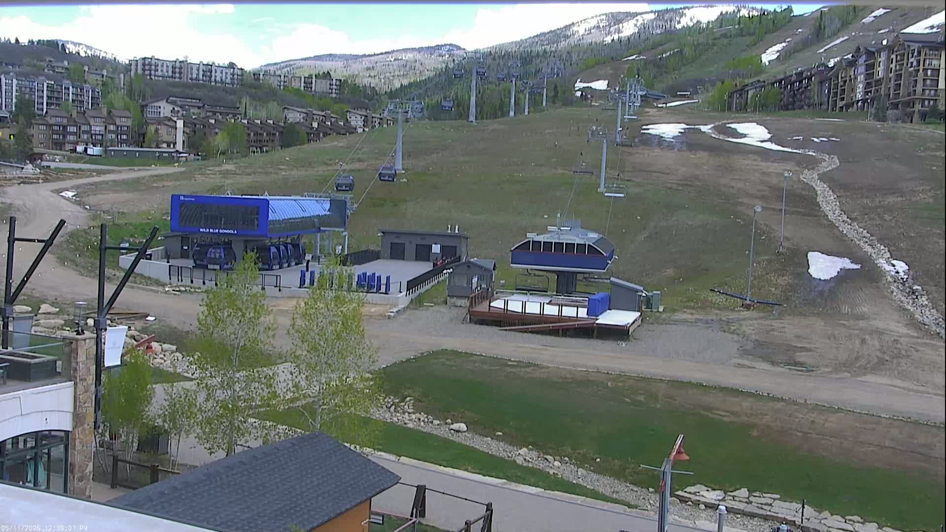 A mostly clear day reveals a ski resort with chairlifts and gondola stations, brown grassy hills, and buildings in the background.