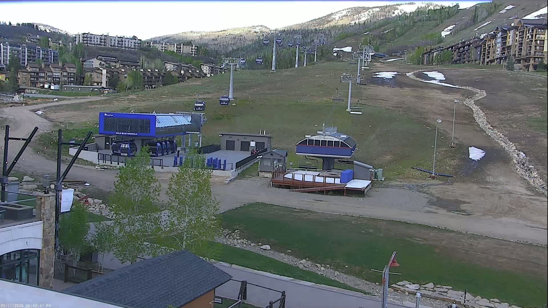 A ski resort in the mountains on a mostly clear day shows several chairlifts and gondola stations, with patches of remaining snow on the ground.