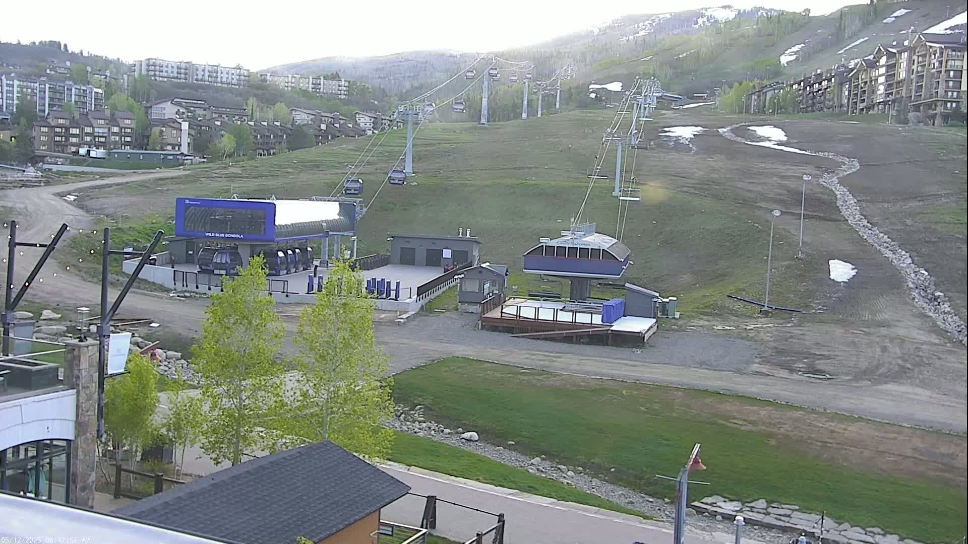 A ski lift base area with several buildings and chairlifts against a hillside with patchy grass and some remaining snow under a mostly clear sky.