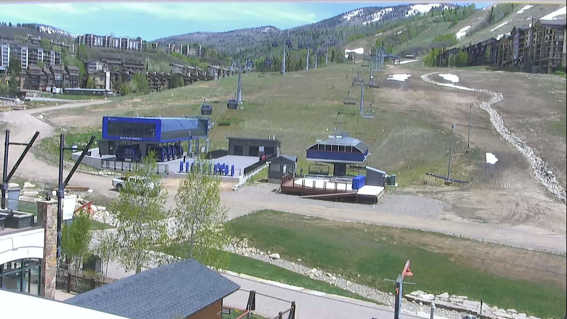 A mostly sunny view of a ski resort base area with several chairlifts, buildings, and a partially snow-covered hillside in the background.