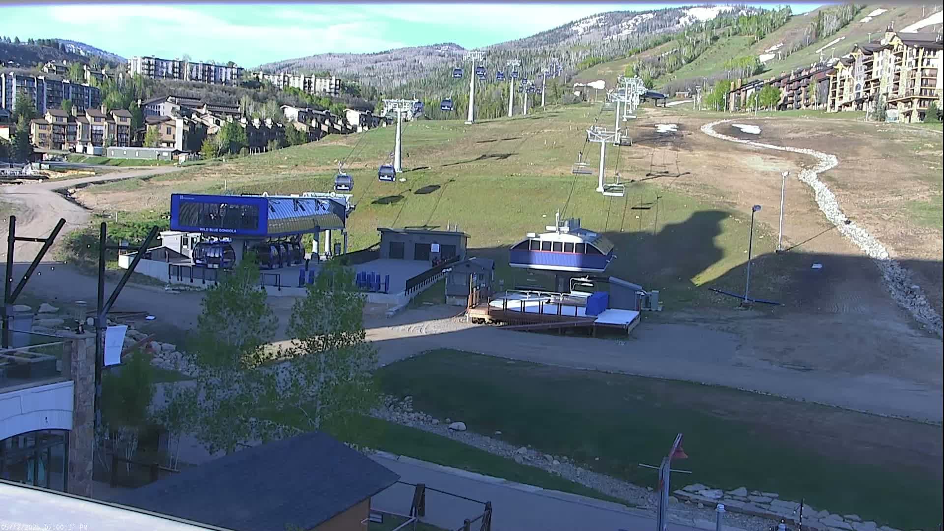 Two ski lift stations are situated on a grassy slope below a mountainside development, under a mostly sunny sky.