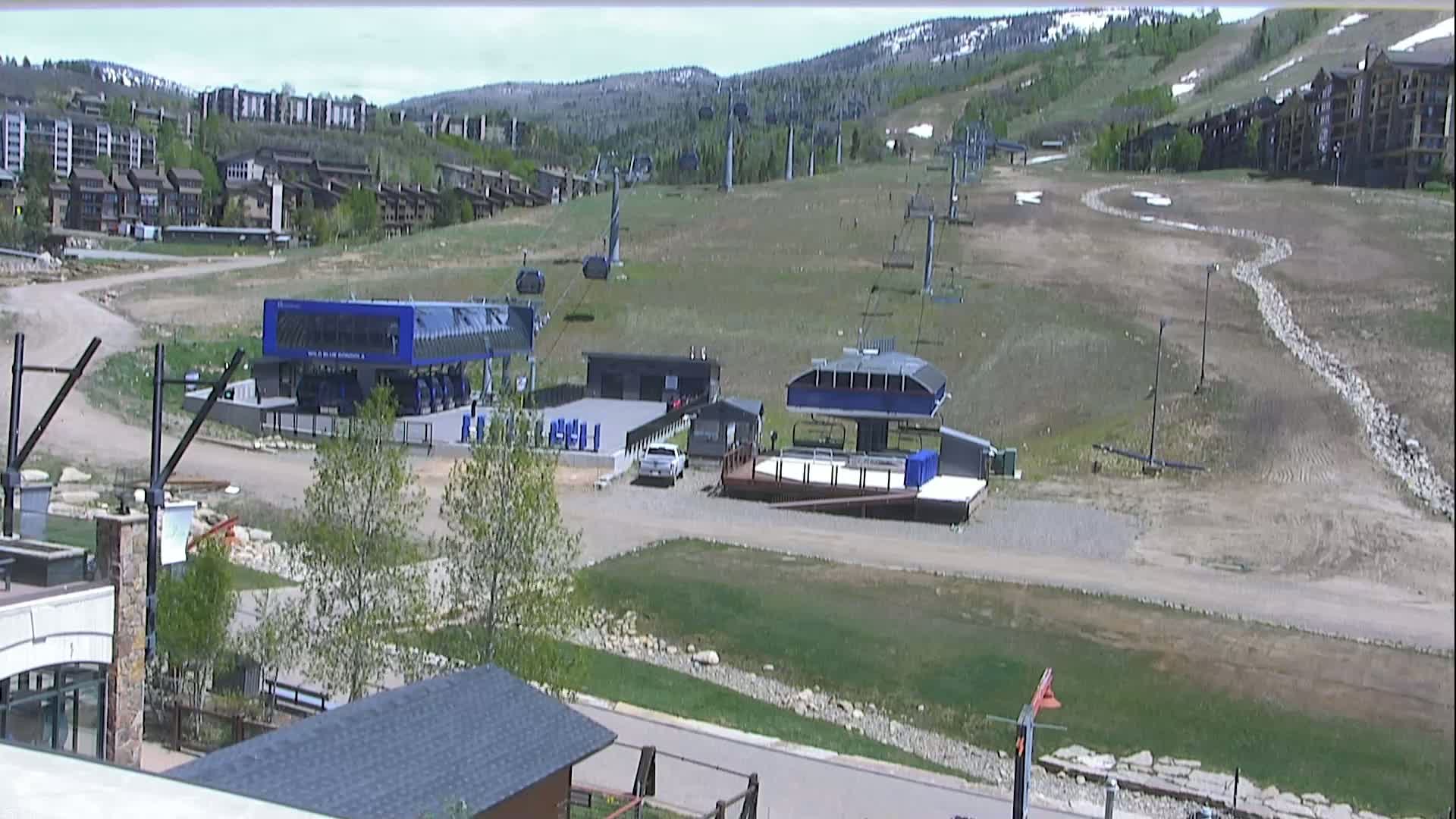 A mostly sunny day shows a ski resort with several chairlifts, buildings, and a few vehicles in a grassy area against a backdrop of mountains.