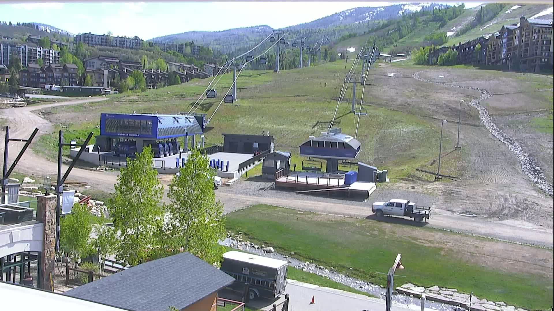 A sunny day at a ski resort shows empty ski lifts, a few buildings, and a truck parked on a mostly barren hillside with some green grass and a small stream.