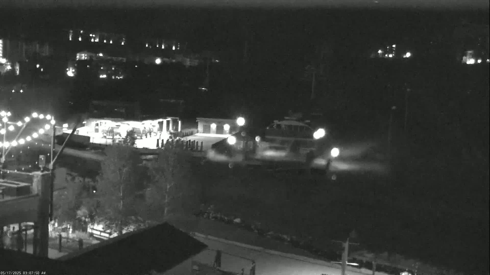 At night, two brightly lit vehicles are parked near a building, with a string of lights visible in the upper left corner and some trees in the mid-ground.