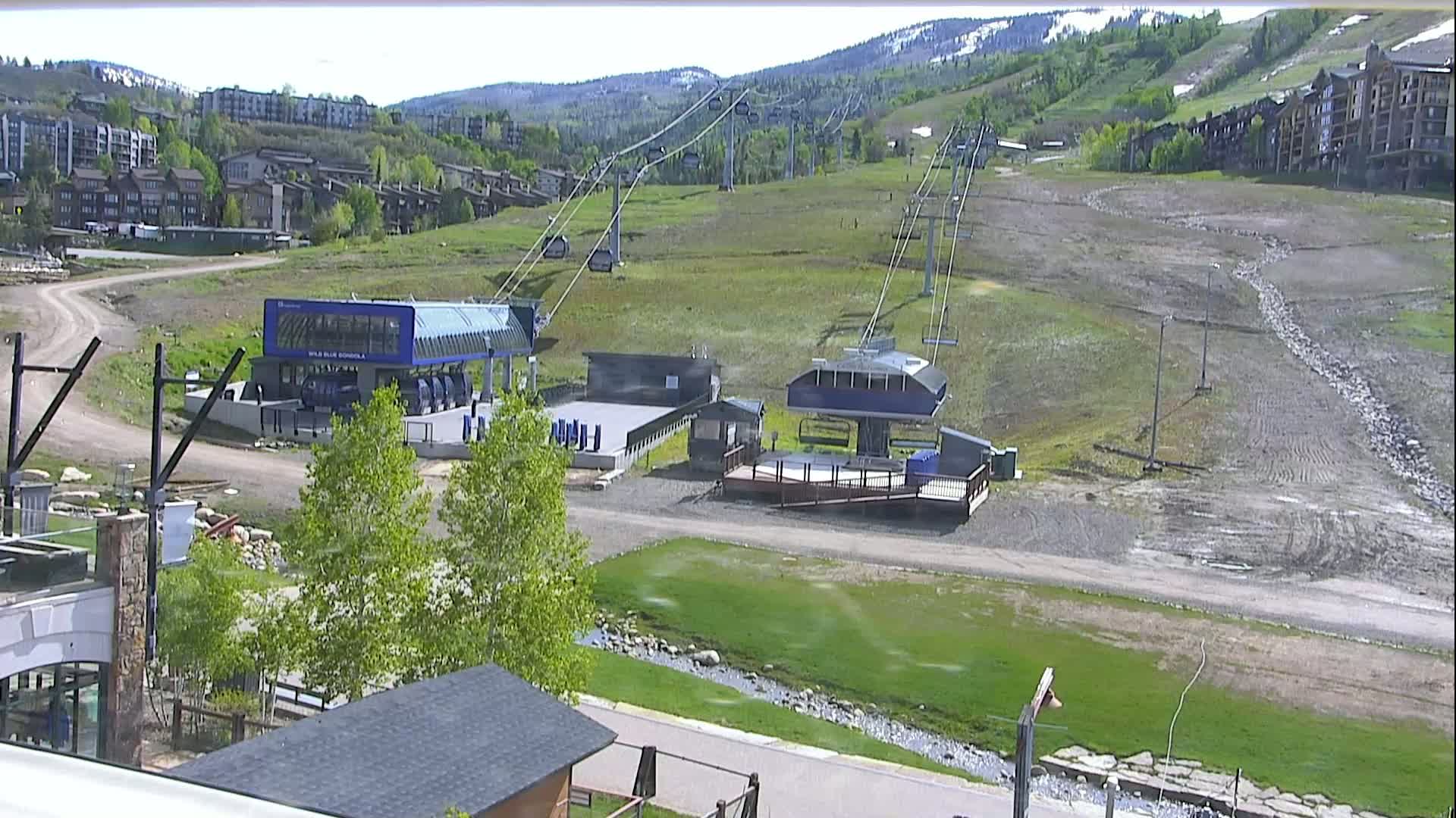A ski lift base station and gondola terminal sit on a grassy hillside, with buildings and a mountain range visible in the background under a clear sky.