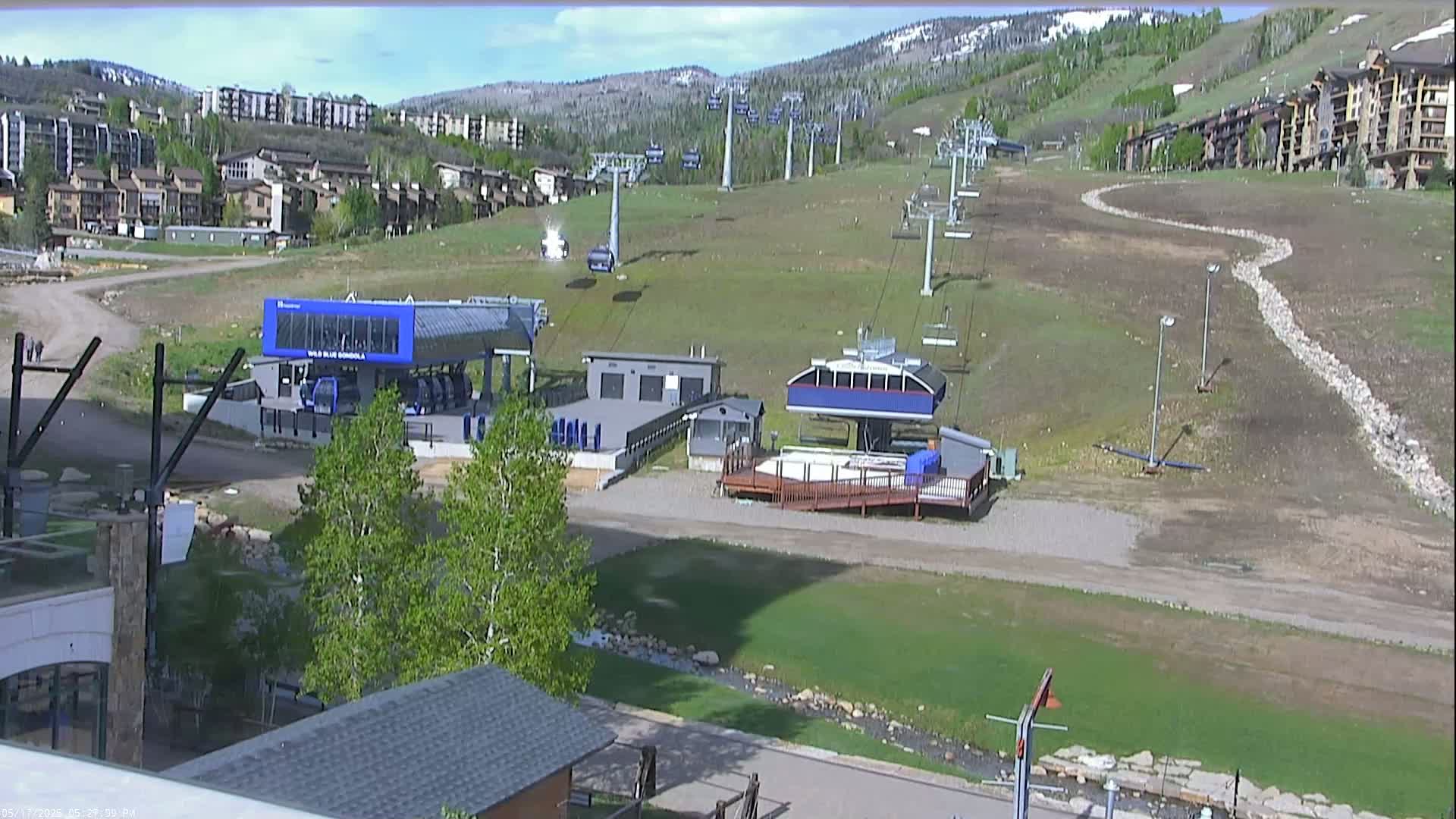 A sunny view of a ski resort base area with chairlifts, buildings, and grassy slopes.