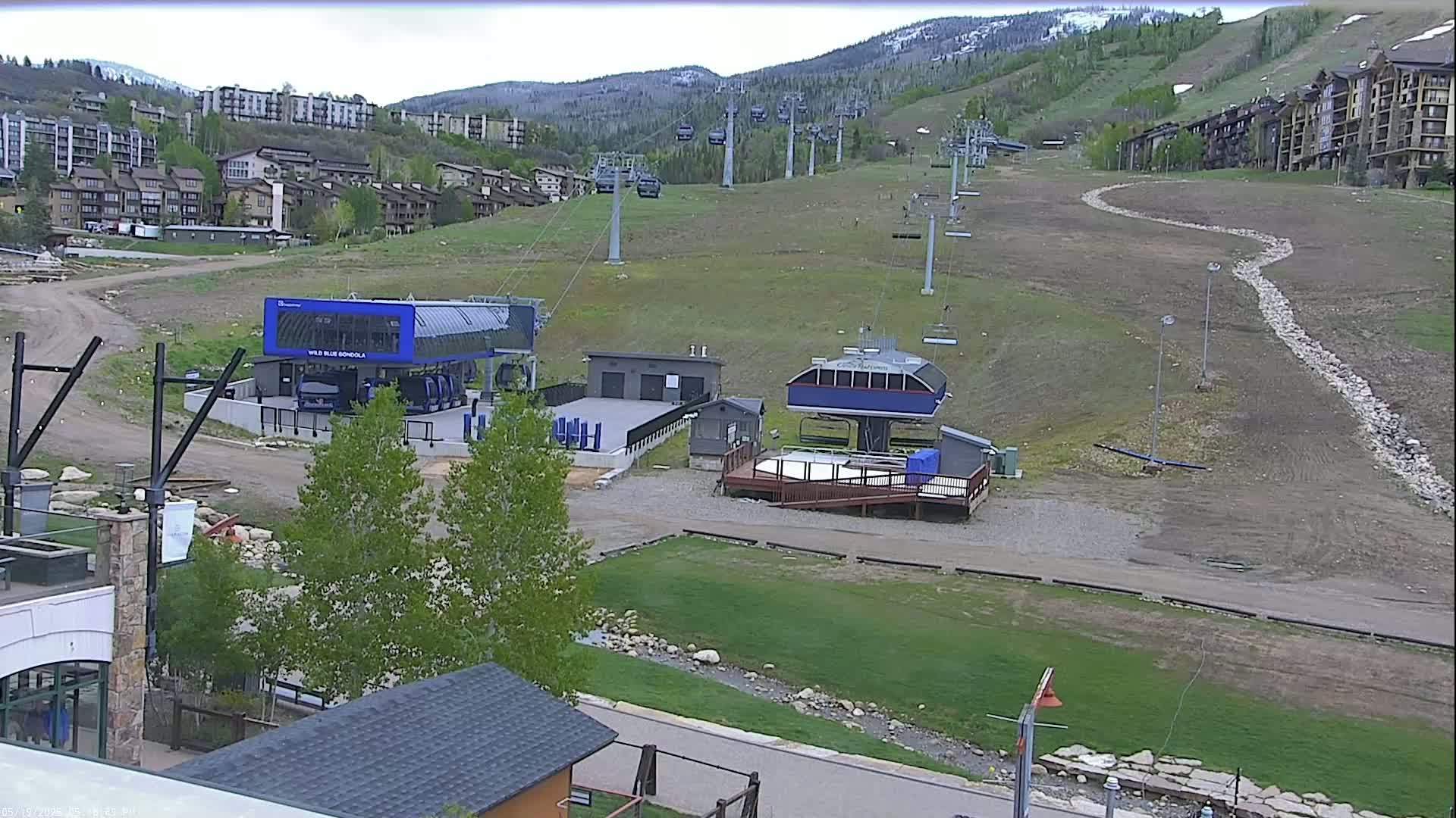 A mostly cloudy day shows a ski resort with several chairlifts and gondola stations, nestled among buildings and green hillsides.