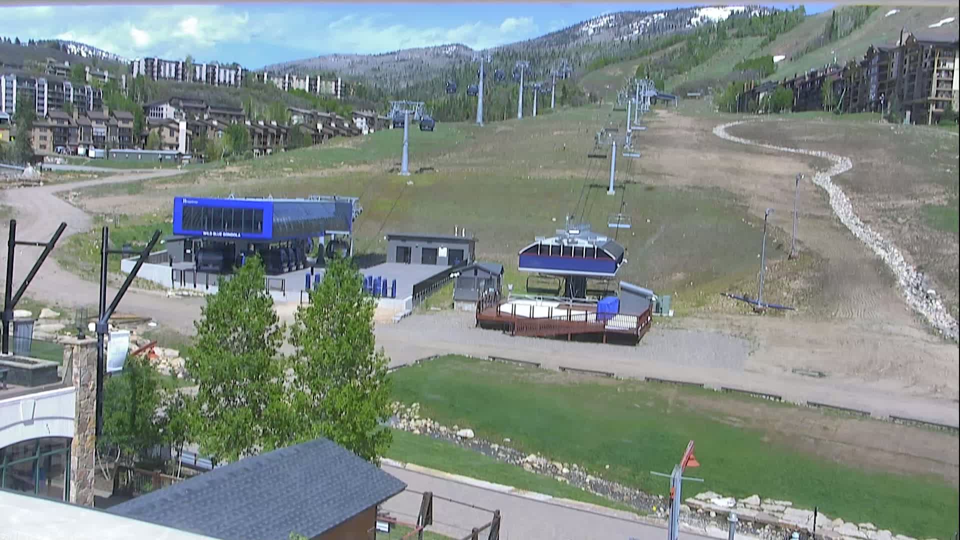 A mostly sunny day reveals a ski resort with chairlifts and base buildings, some green grass, and brown, barren patches of land on the hillside.