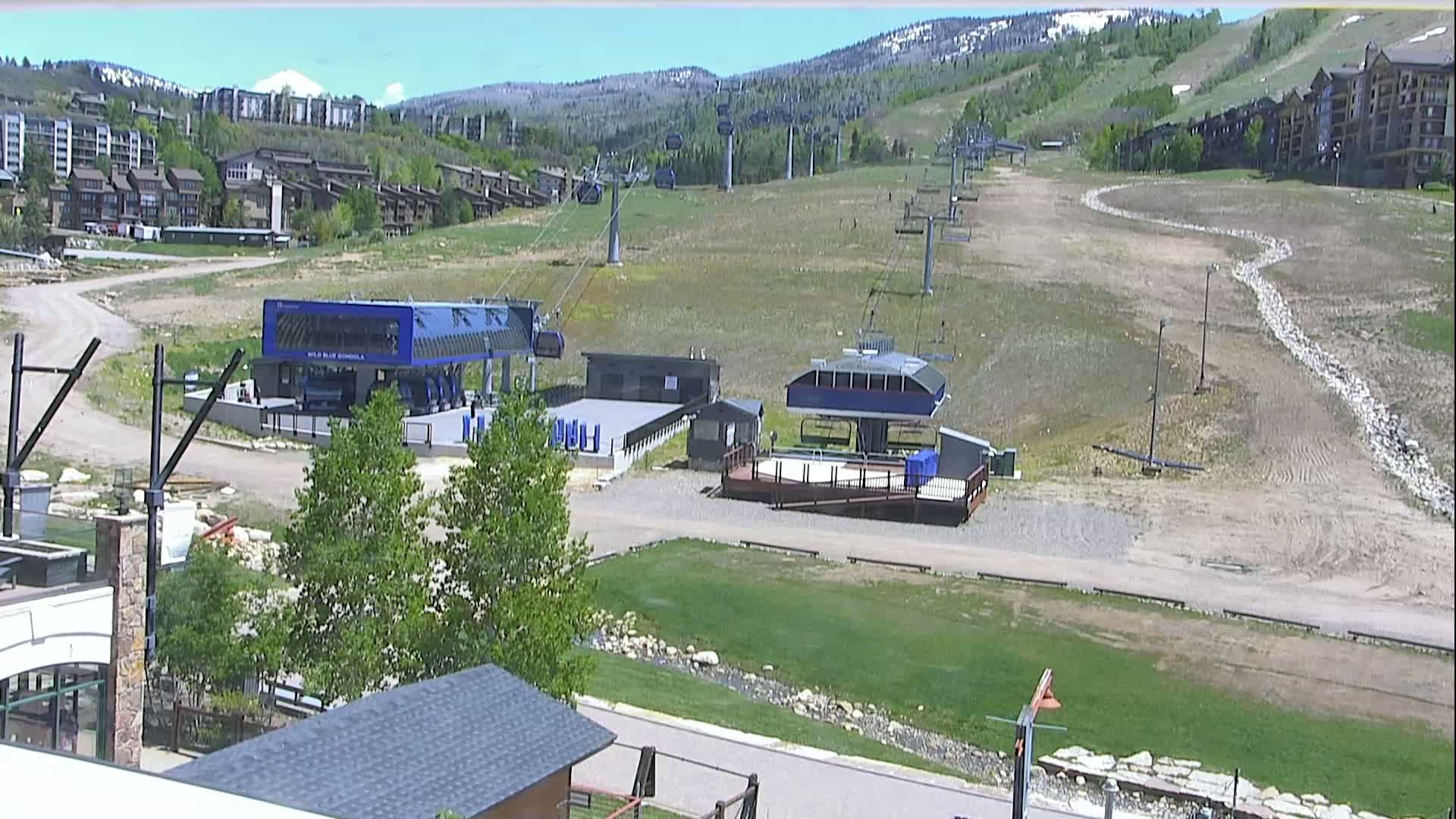 A sunny view of a ski resort in summer, showing two chairlift stations and a grassy hill with ski runs visible in the background.