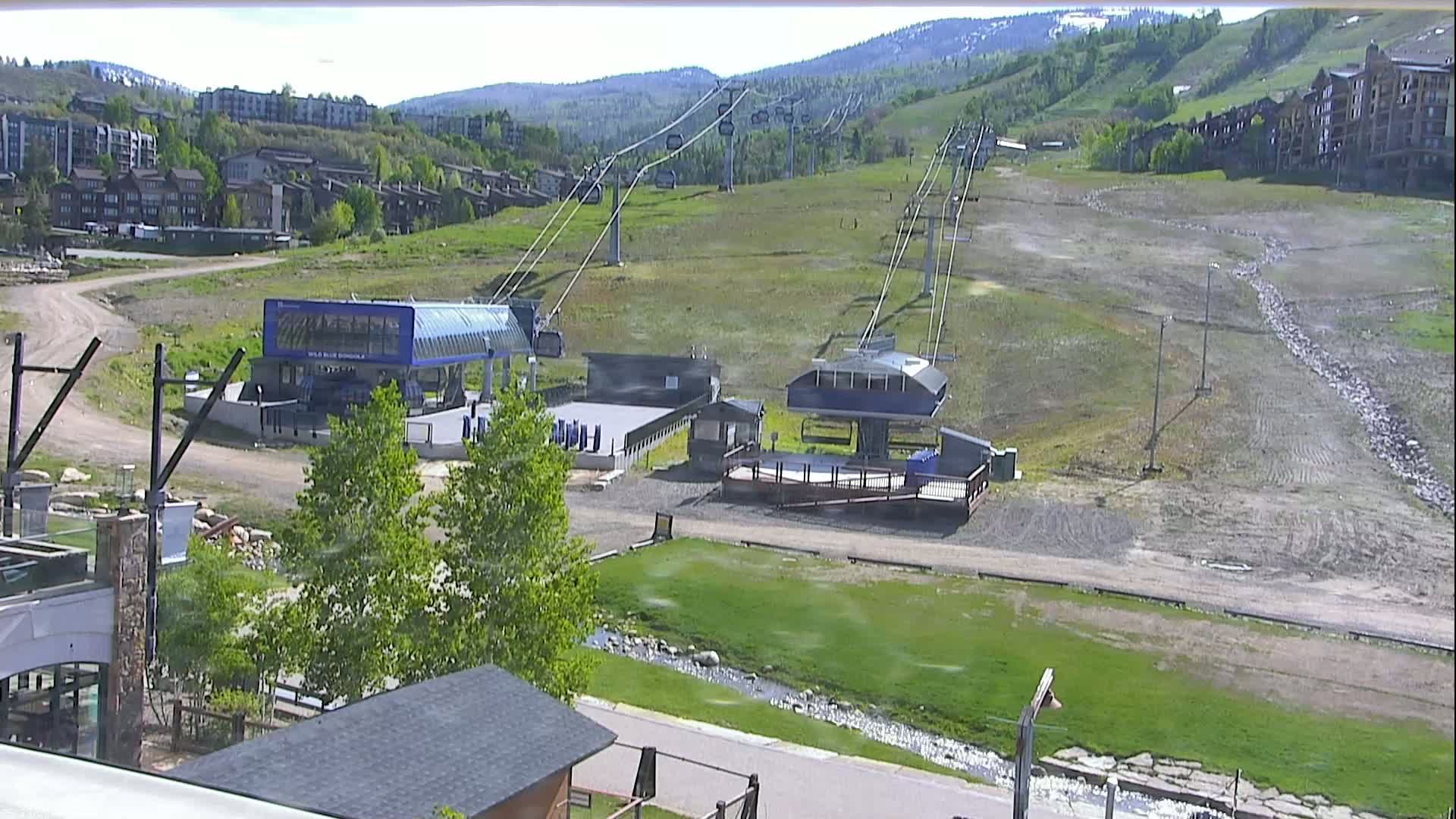 A sunny view of a ski resort in the summer, showing two ski lifts and surrounding buildings, with a small stream running through a grassy area in the foreground.