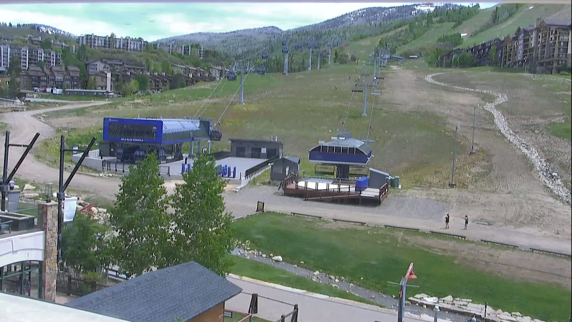 A ski resort base area on a partly cloudy day, showing chairlifts, buildings, and a grassy area with a small stream.