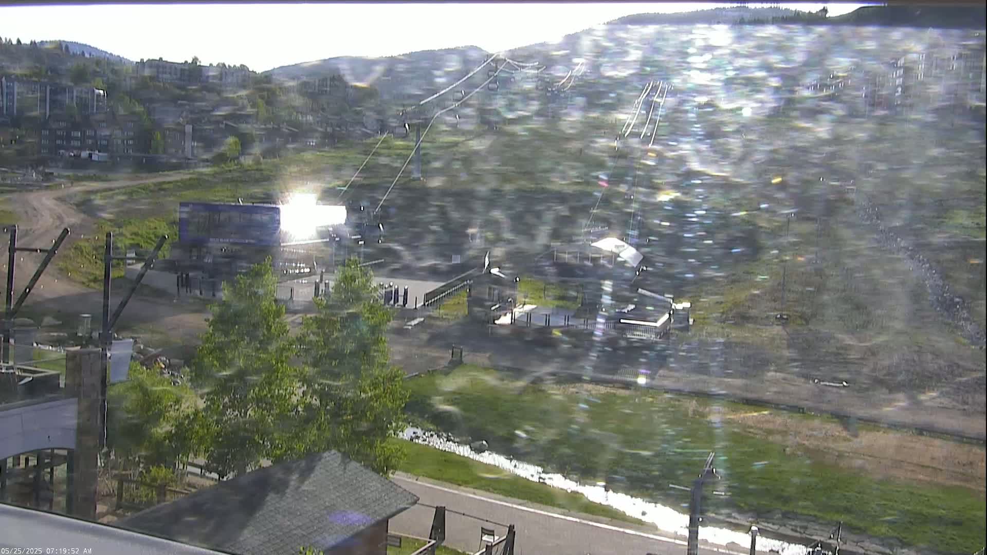 A sunny view of a ski resort base area with a chairlift ascending a mountain, buildings, and a grassy area with a stream.