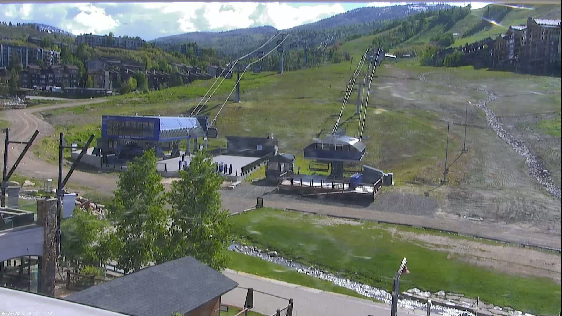 A mostly sunny view of a ski resort base area in summer, showing chairlifts, buildings, and grassy slopes.
