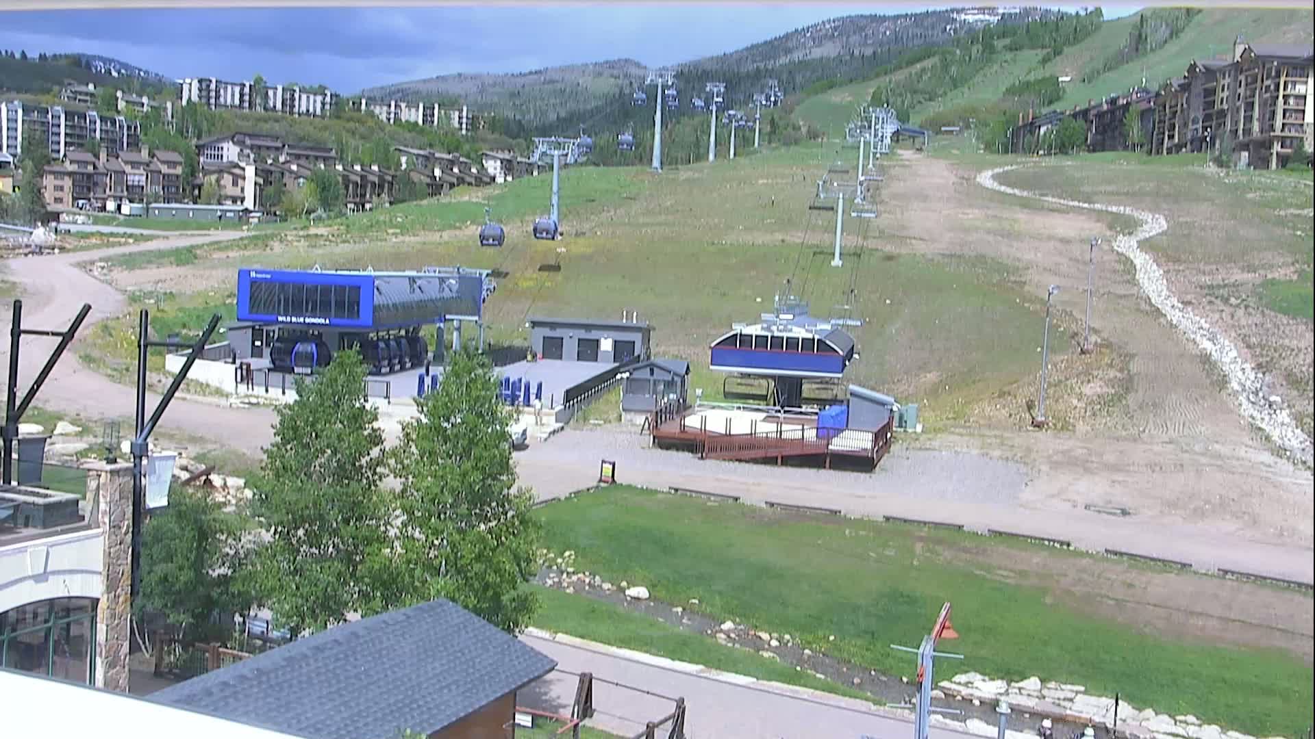 A mostly sunny view of a ski resort in summer, showing several chairlifts and gondola stations, with buildings and green hills in the background.
