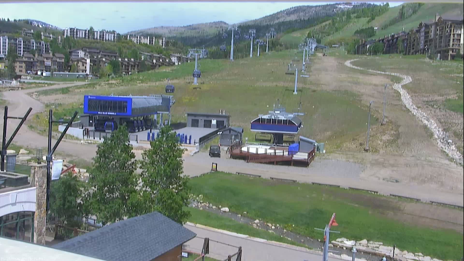 A ski resort base area with several chairlifts, buildings, and a mostly dry, grassy landscape under a partly cloudy sky.