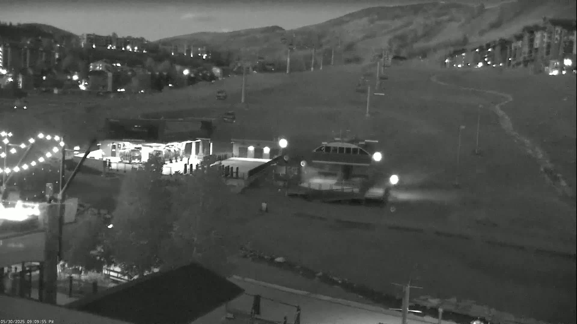A nighttime, infrared view shows a ski resort with a chairlift, buildings, and a road, under clear skies.
