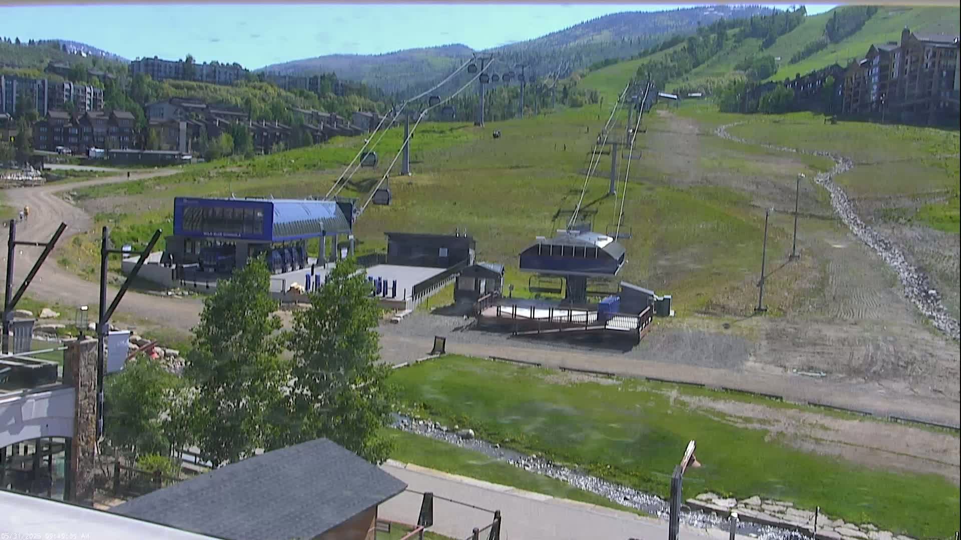 A sunny view of a ski lift and base area, with grassy hills and buildings in the background.
