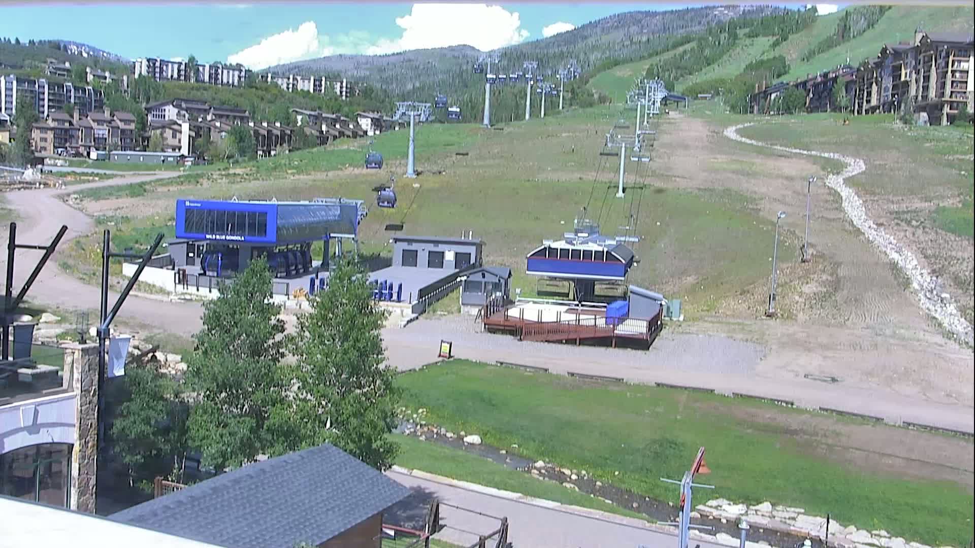 A mostly sunny day reveals a ski resort base area with several chairlifts, buildings, and grassy slopes.