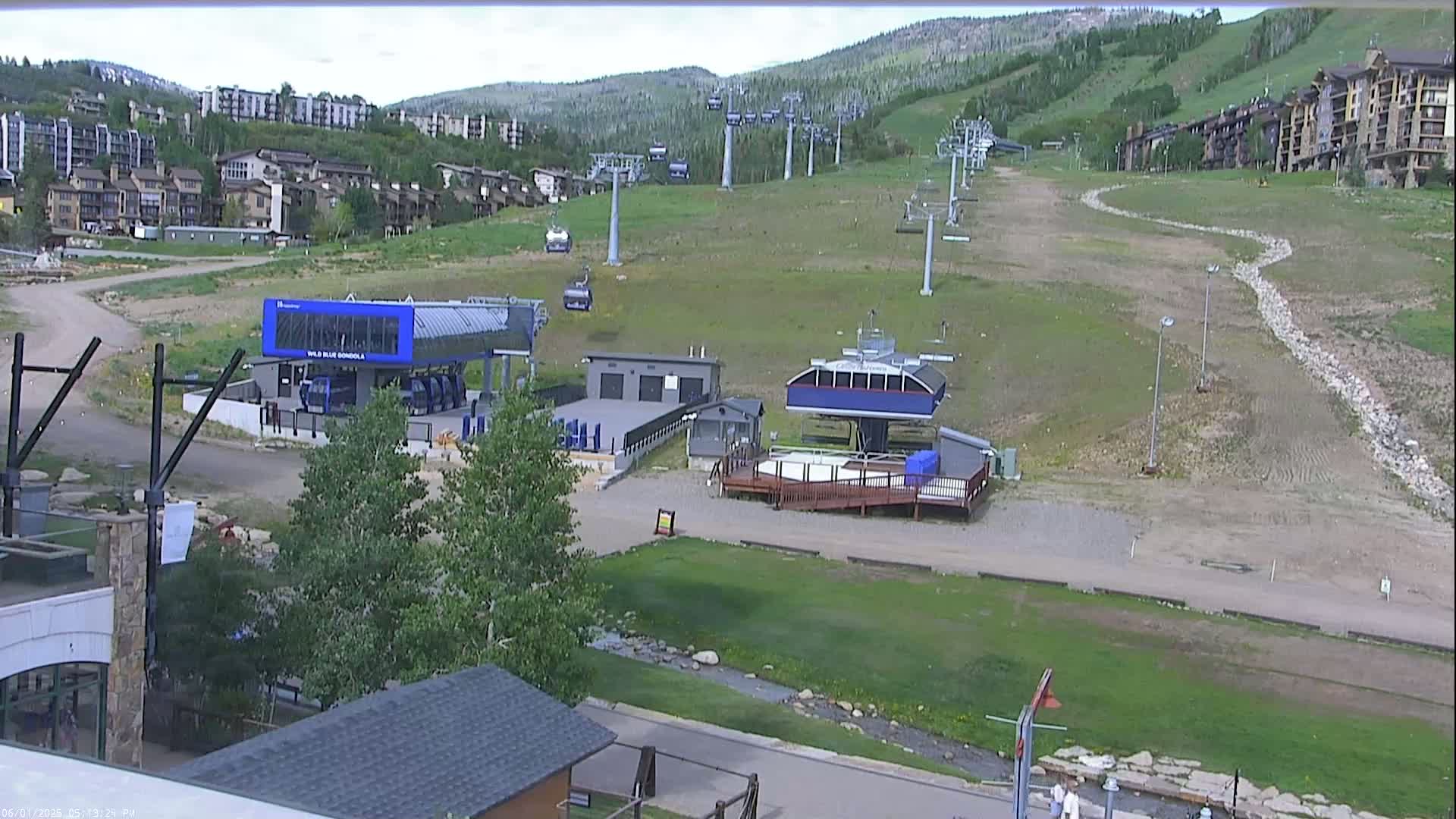 A ski resort in the summer shows several chairlifts and gondola stations with buildings and green grassy areas, mountains in the background under a mostly clear sky.