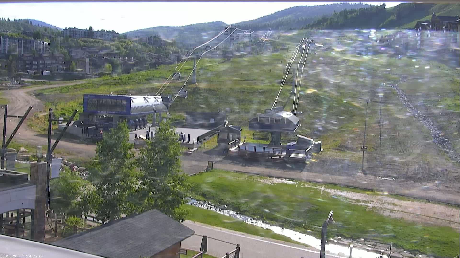A ski lift and its base station are visible on a grassy hillside under a bright, windy sky, with buildings and a small stream in the foreground.