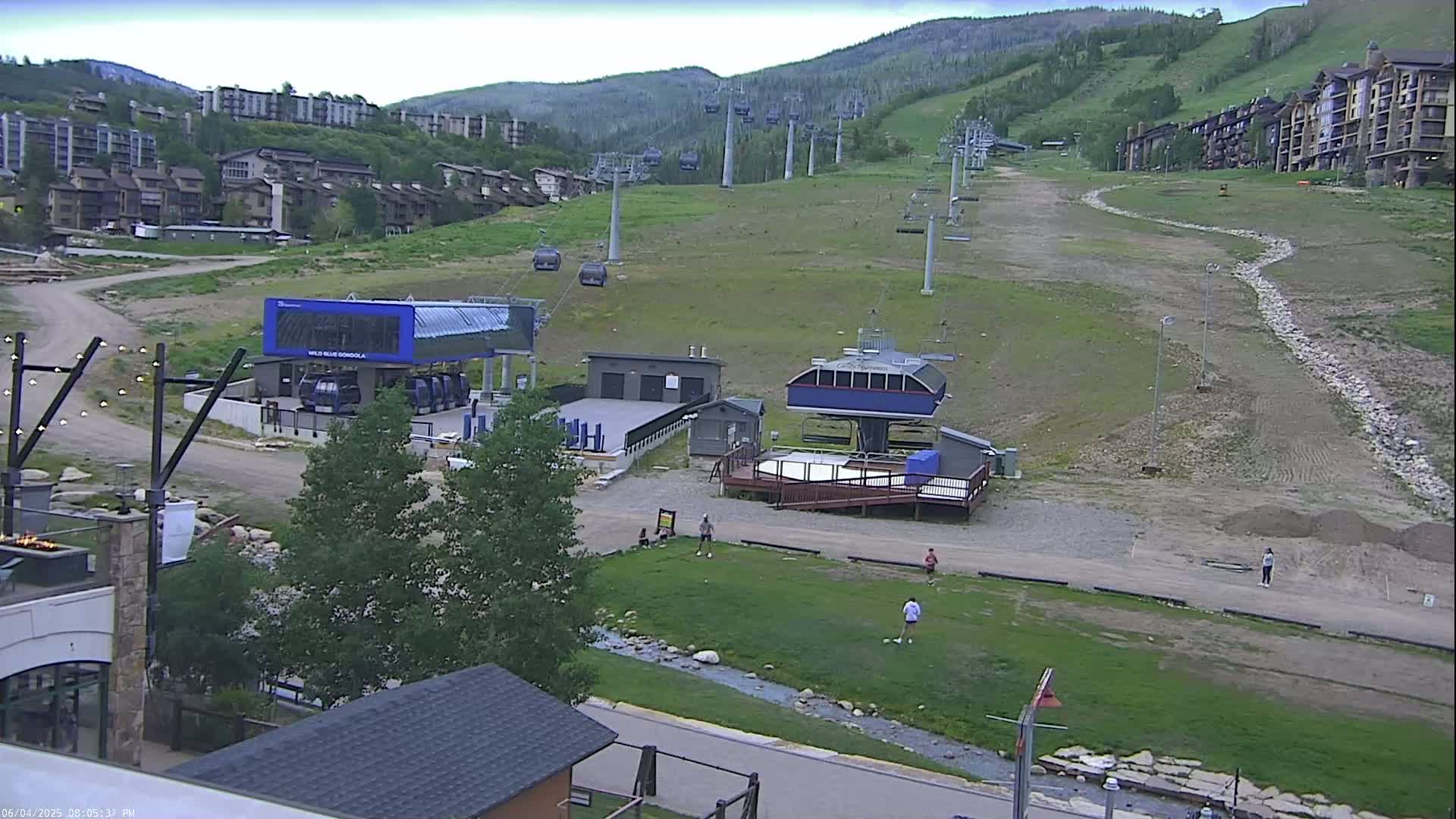 A ski resort in the mountains on an overcast day shows several chairlifts and buildings, with a few people visible in a grassy area near the base of a lift.
