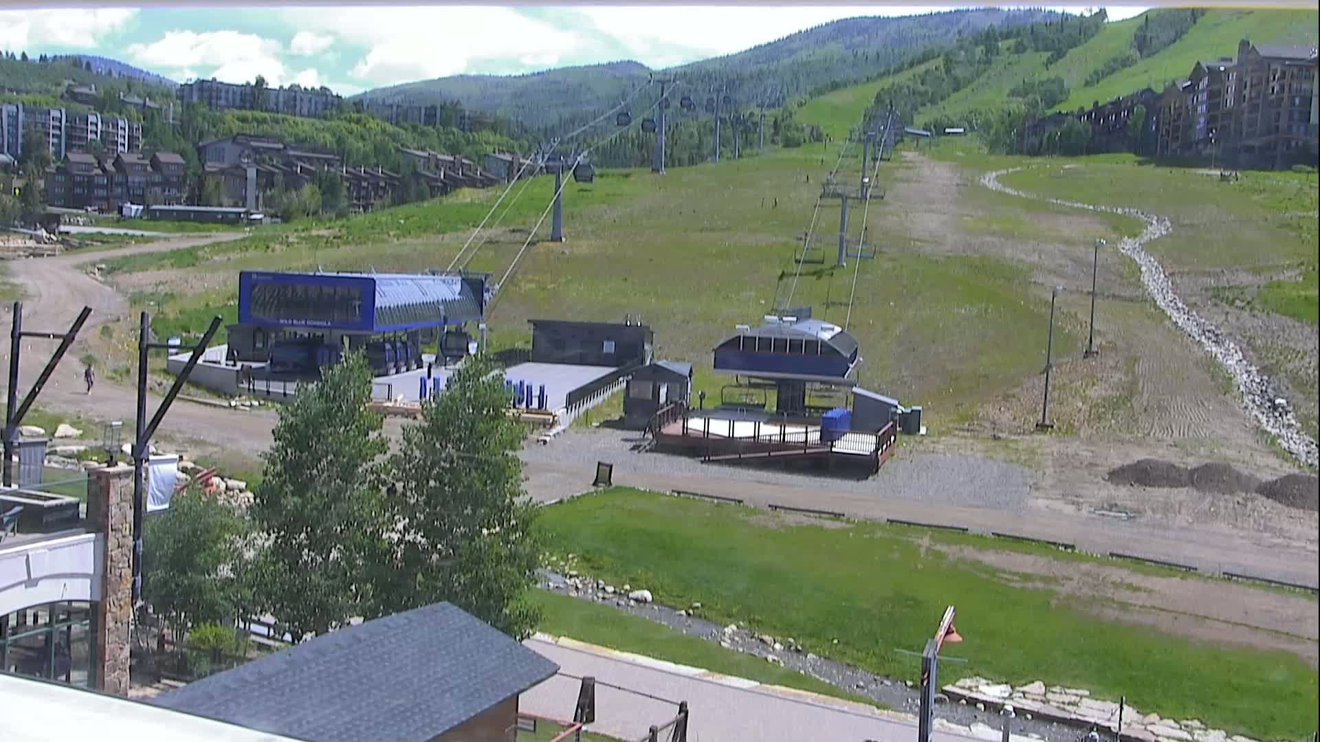 A ski lift base area with two chairlift stations, surrounded by grassy hills and residential buildings under a partly cloudy sky.