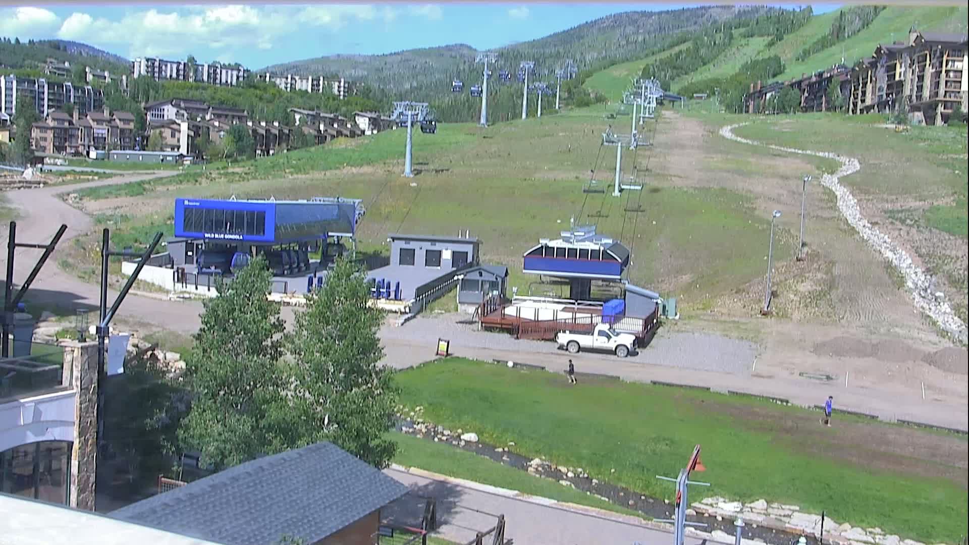 A mostly sunny view of a ski resort in summer shows several chairlifts, buildings, and a pickup truck, with a small stream visible in the foreground.