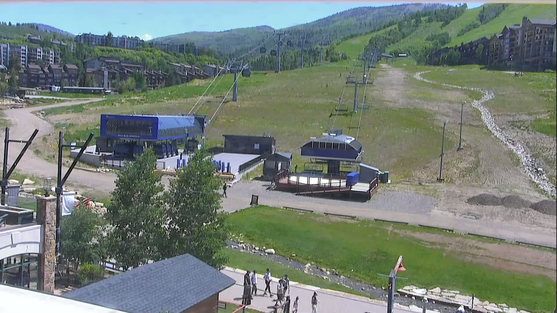 A sunny day reveals a ski resort with two chairlift stations, surrounding buildings, and people walking on a paved path.
