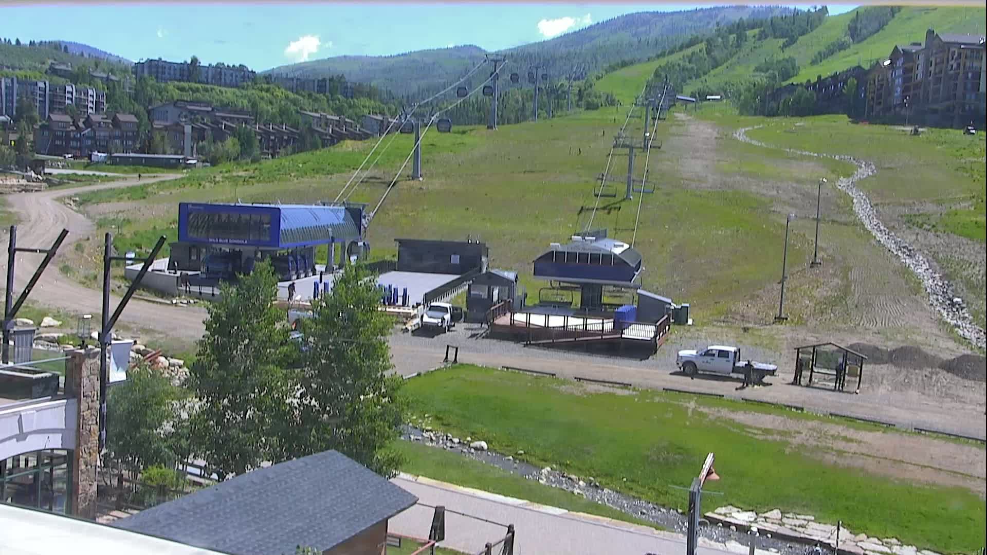 A ski lift base area with two lift stations, several vehicles, and some people is visible on a sunny day against a backdrop of green hills and residential buildings.