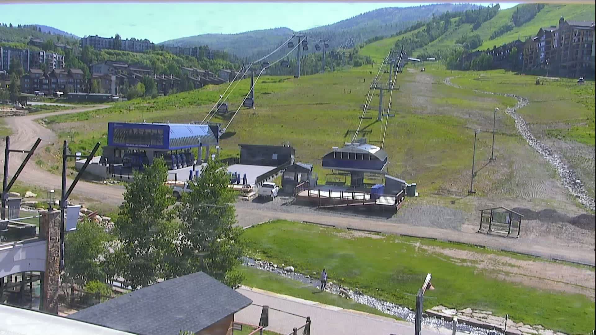 A sunny day reveals a ski resort's chairlifts and base area, with green grass and buildings in the background.