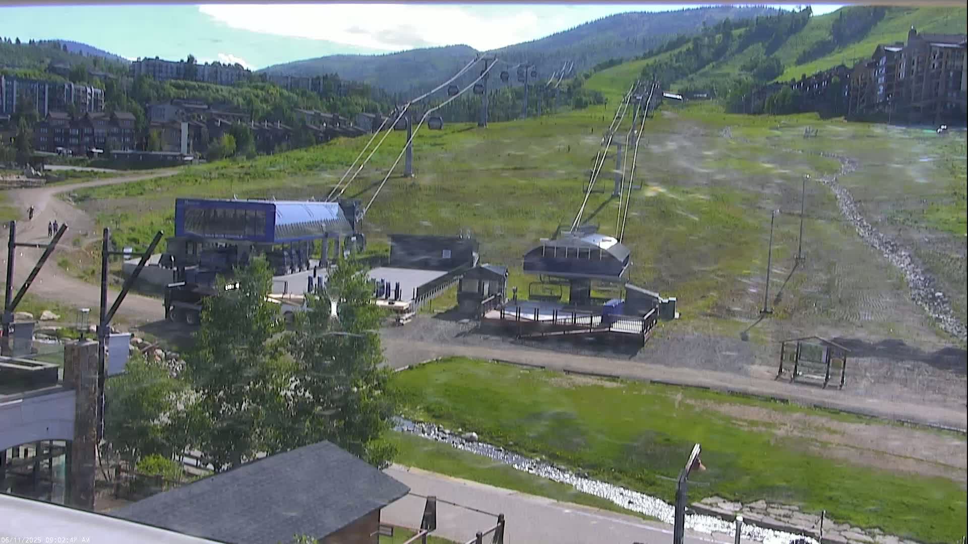 A sunny view of a ski resort in summer, showing two chairlift stations, a grassy hill, and buildings in the background.