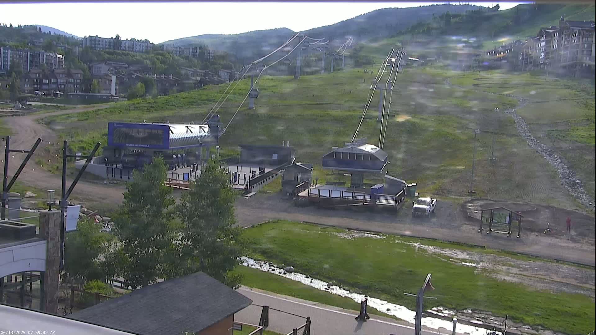 A ski lift base area with two chairlifts extending up a grassy mountainside on an overcast day with light rain.