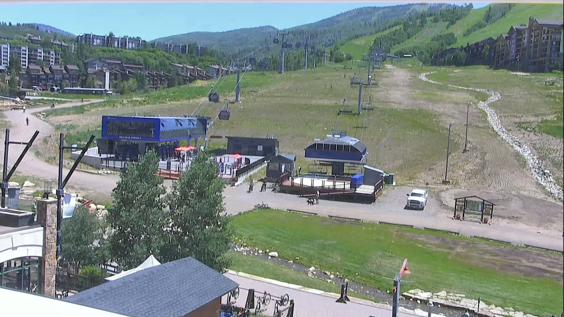 A sunny, slightly hazy view of a mountainside resort area with buildings, roads, and green spaces, including a small stream.
