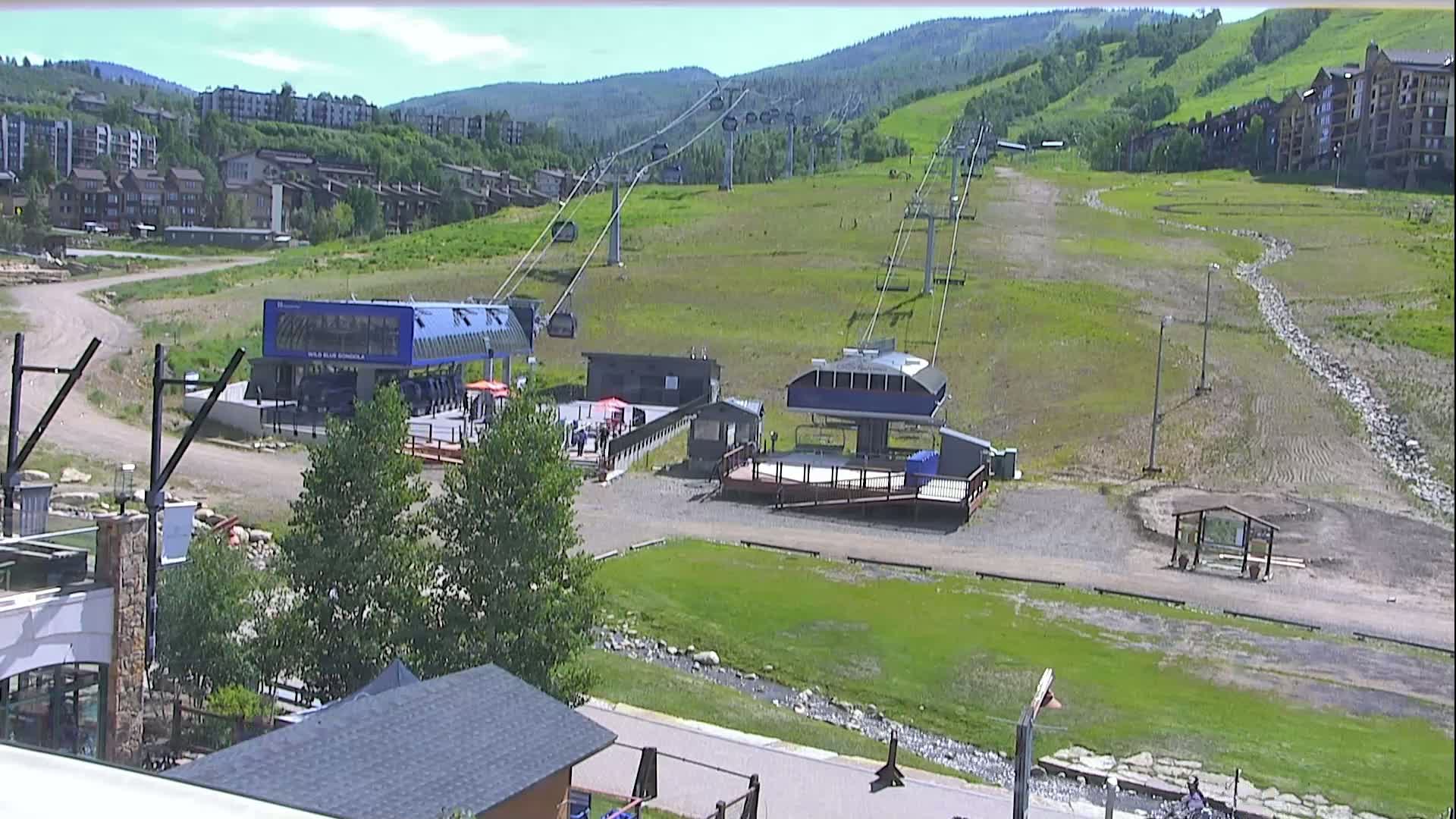 Two ski lift stations sit on a grassy hill in front of residential buildings, under a mostly sunny sky.