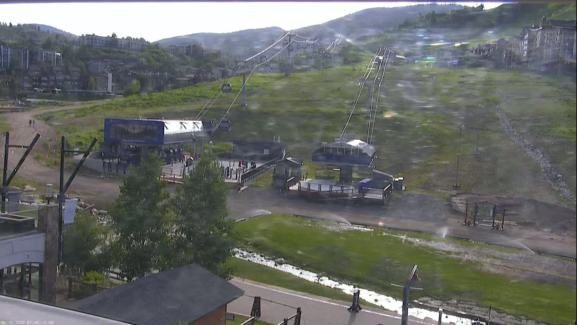 A ski lift base area with two lift stations is shown on a hazy, windy day, with green grass and a small stream visible in the foreground.