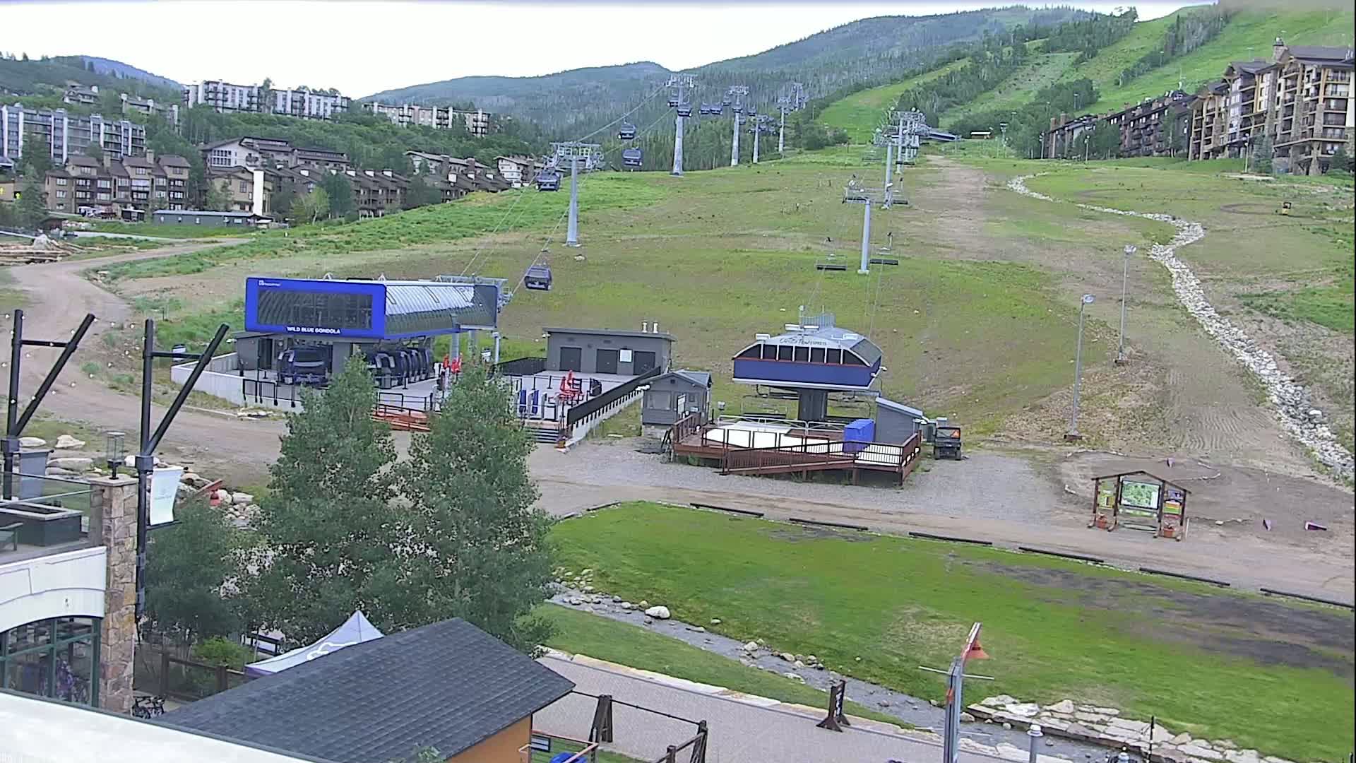 A ski resort's base area with several chairlifts and gondola stations is visible, under partly cloudy skies, with buildings and grassy hills in the background.