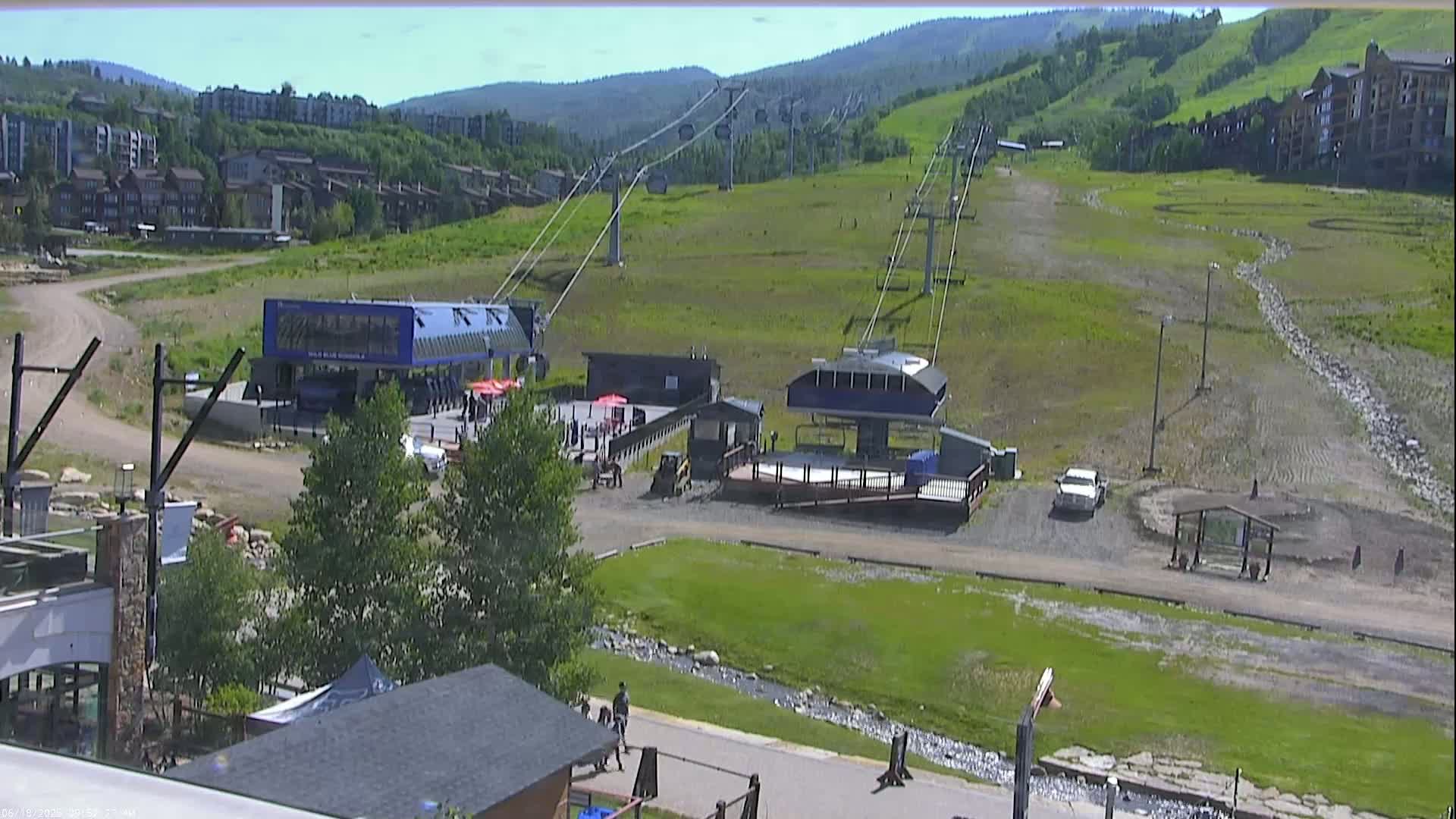 A sunny view of a ski resort's base area shows two chairlift stations, grassy slopes, and buildings, with a small stream running through the lower portion.