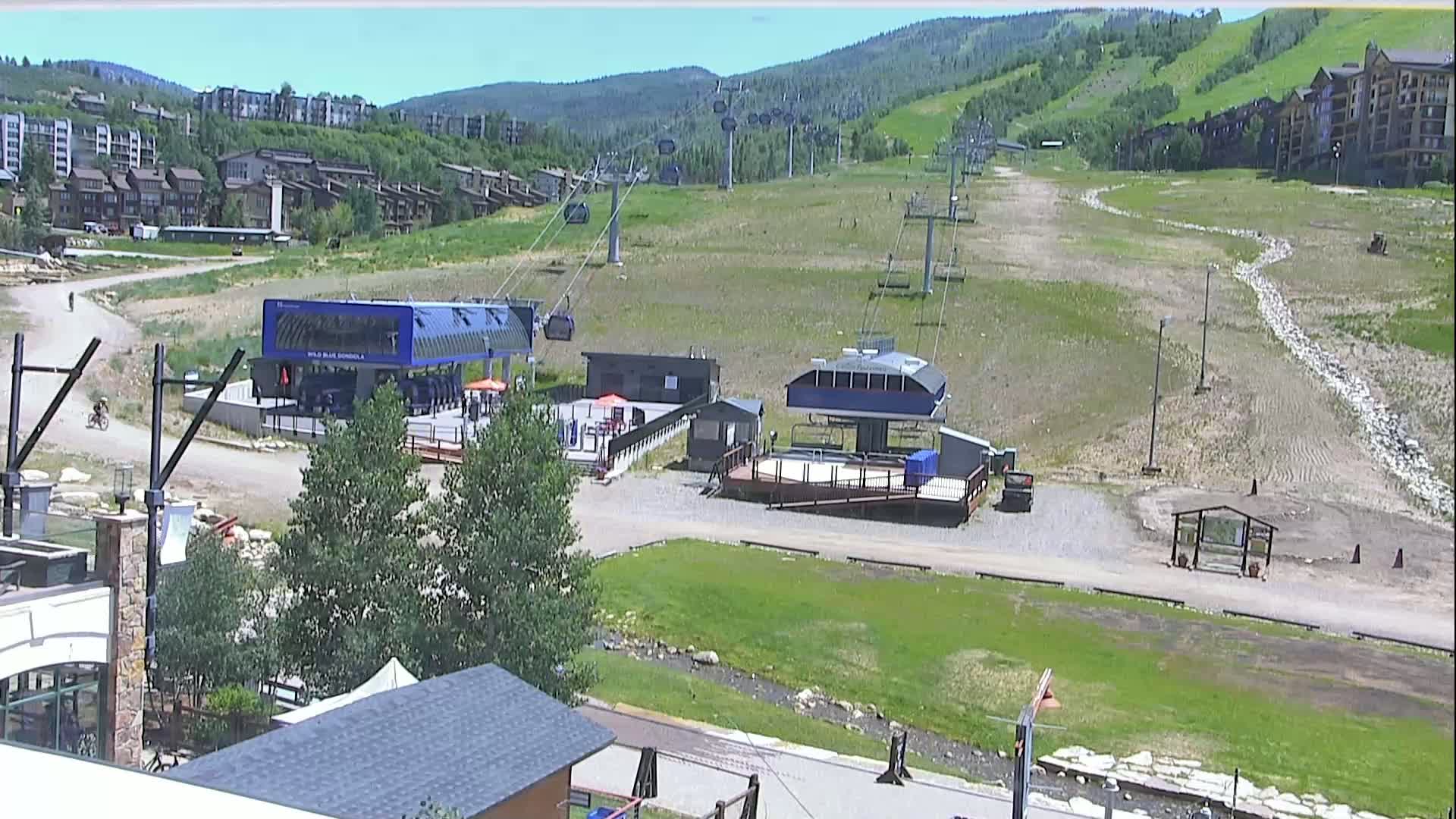 A ski lift base area with two lift stations, a grassy hillside, and buildings in the background on a sunny day.