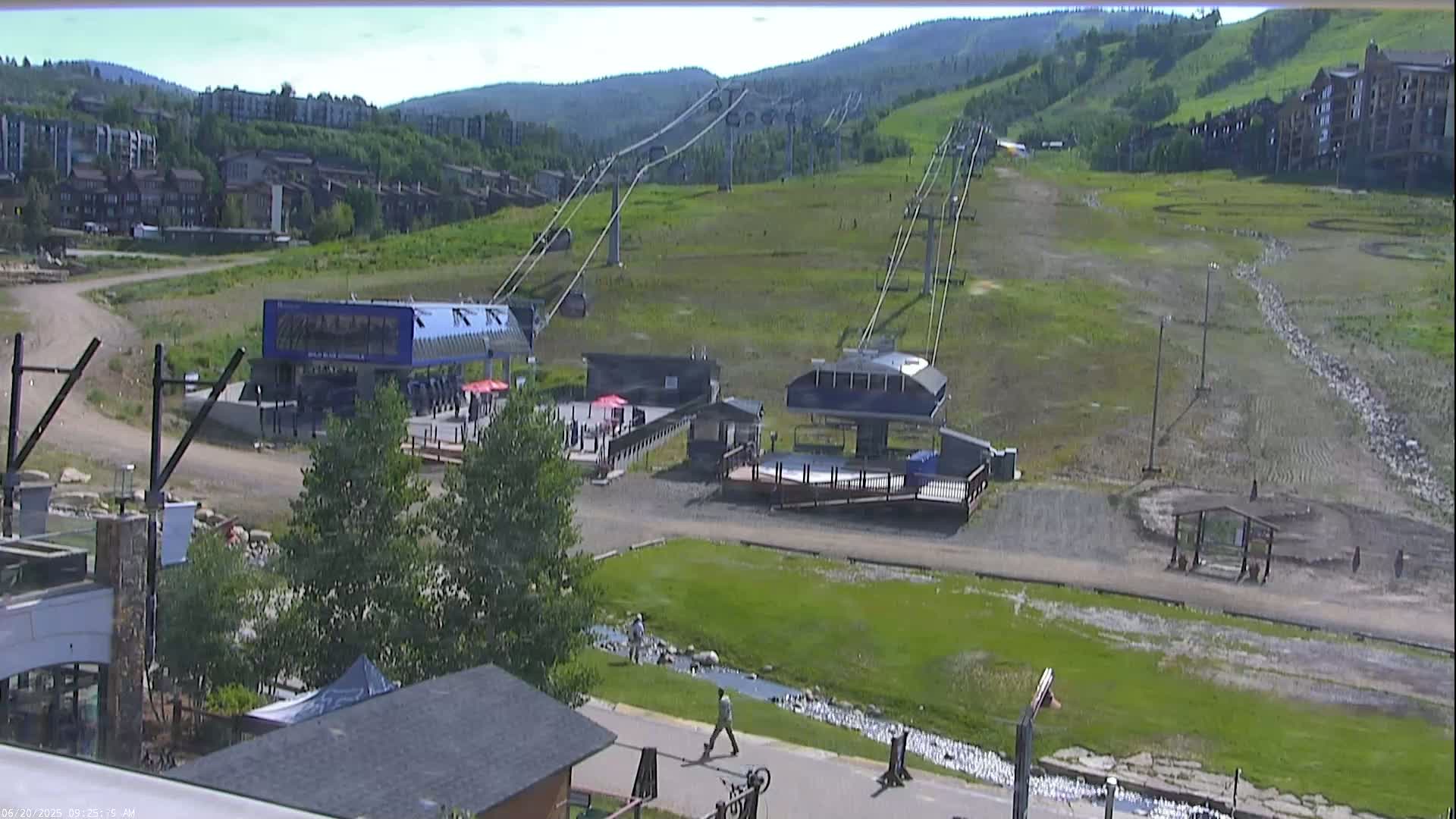 A ski lift base area with two chairlift stations, buildings, and a grassy hillside visible under a sunny sky.
