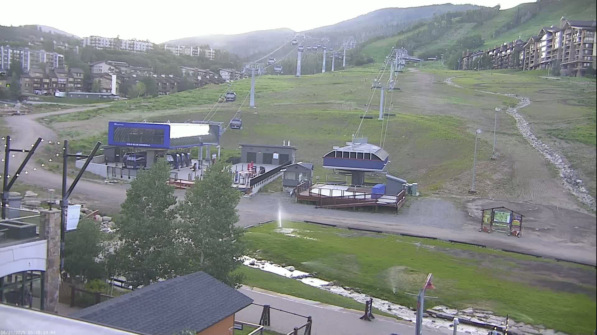 A ski lift base area with several buildings and chairlifts ascending a grassy hill under an overcast sky.
