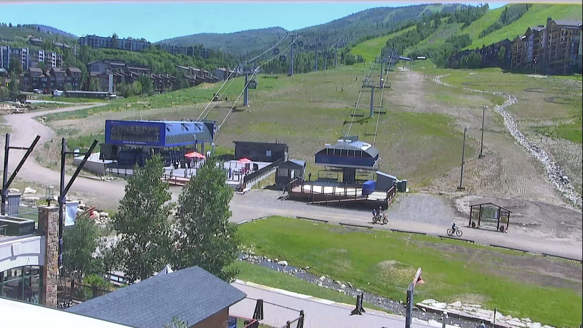 A mountainside ski resort in summer, showing two chairlift stations, several buildings, and people biking on a path, under sunny skies.