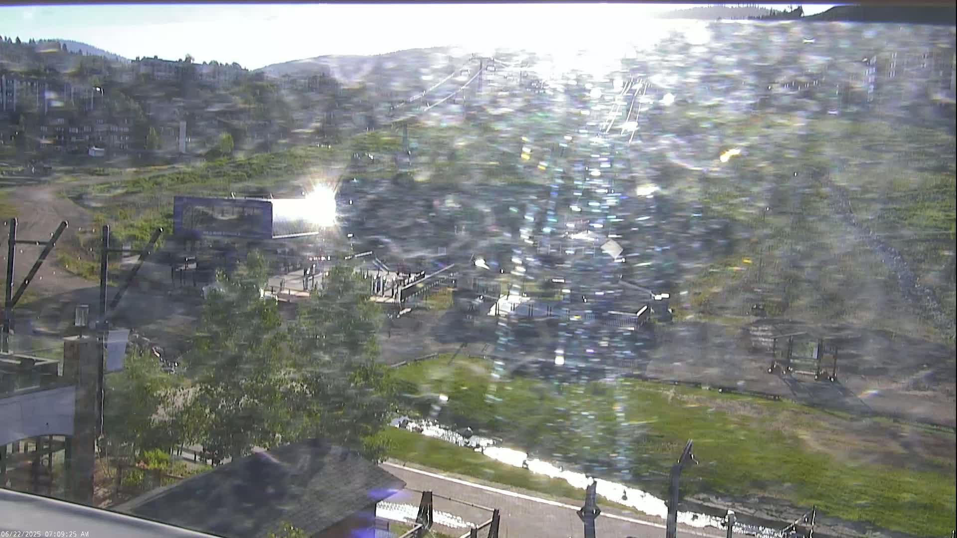 A sunny, somewhat hazy view overlooks a mountain resort area with a chairlift, buildings, and a small stream.