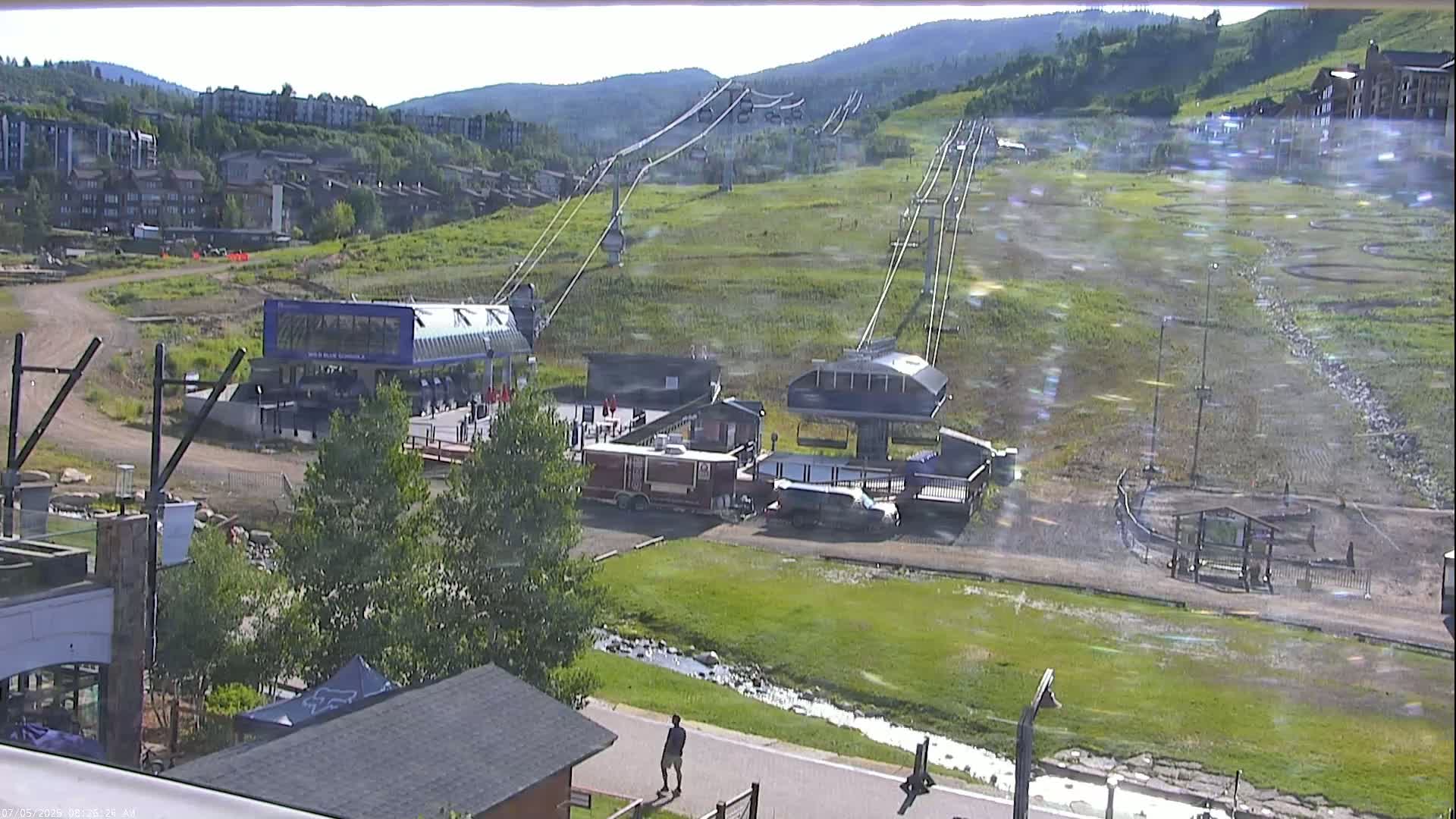 A chairlift ascends a grassy mountainside on a clear, sunny day, with buildings and a small stream visible below.