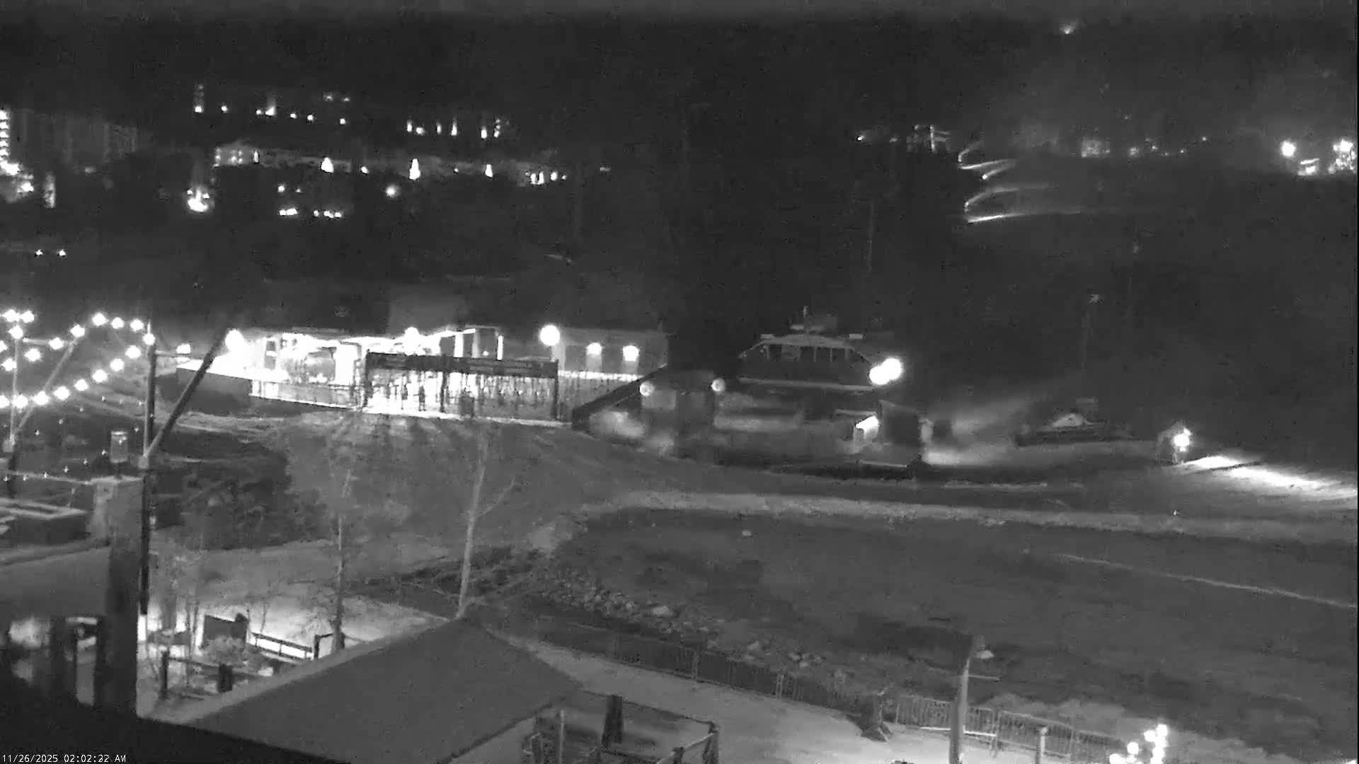 A nighttime, black-and-white scene depicts a snowy ski resort featuring illuminated buildings, a ski lift station, and a snow groomer on the slopes, with what appears to be active snowmaking under cold and clear conditions.